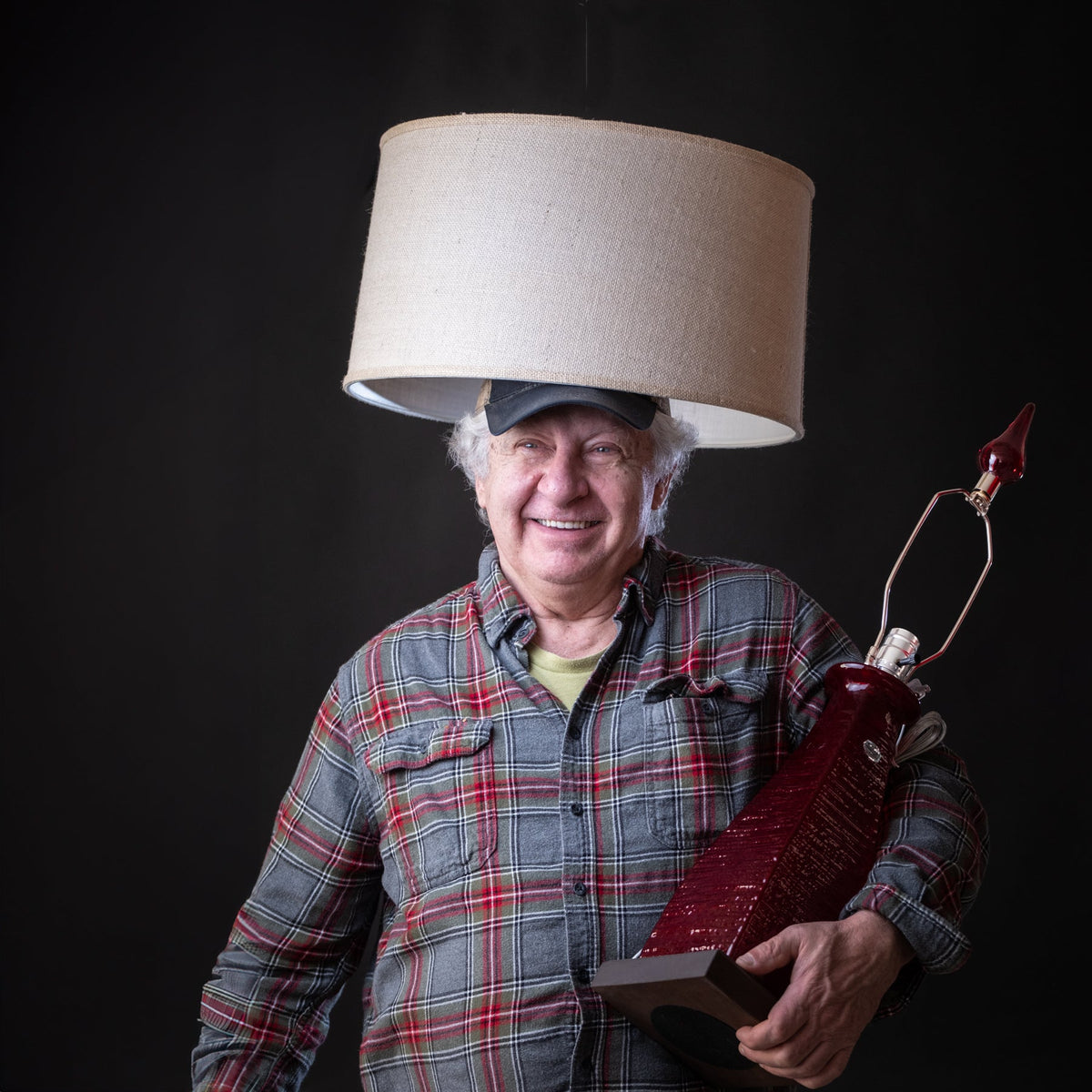 Man wearing a lampshade on his head holding a red bottle against a black background