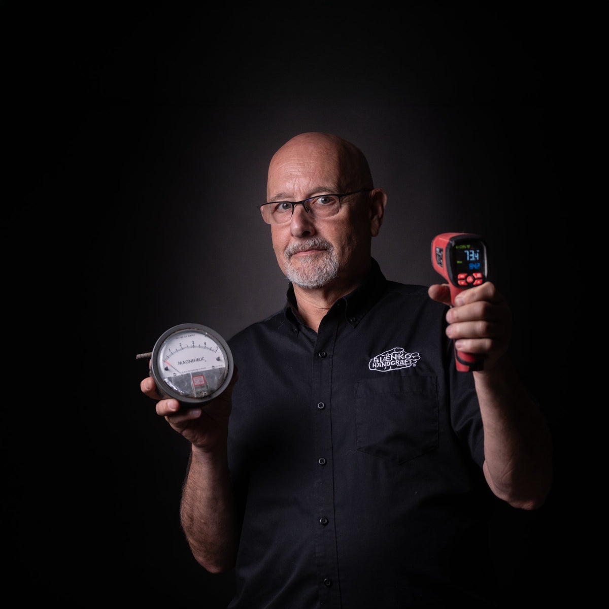 A man wearing glasses and a black button-up shirt holds a round analog gauge in one hand and a red digital thermometer in the other against a dark studio background.