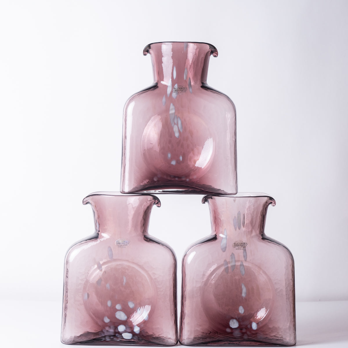 Three translucent dusty pink glass bottles with a rounded rectangular shape and flared necks, stacked in a pyramid arrangement on a clean white background.