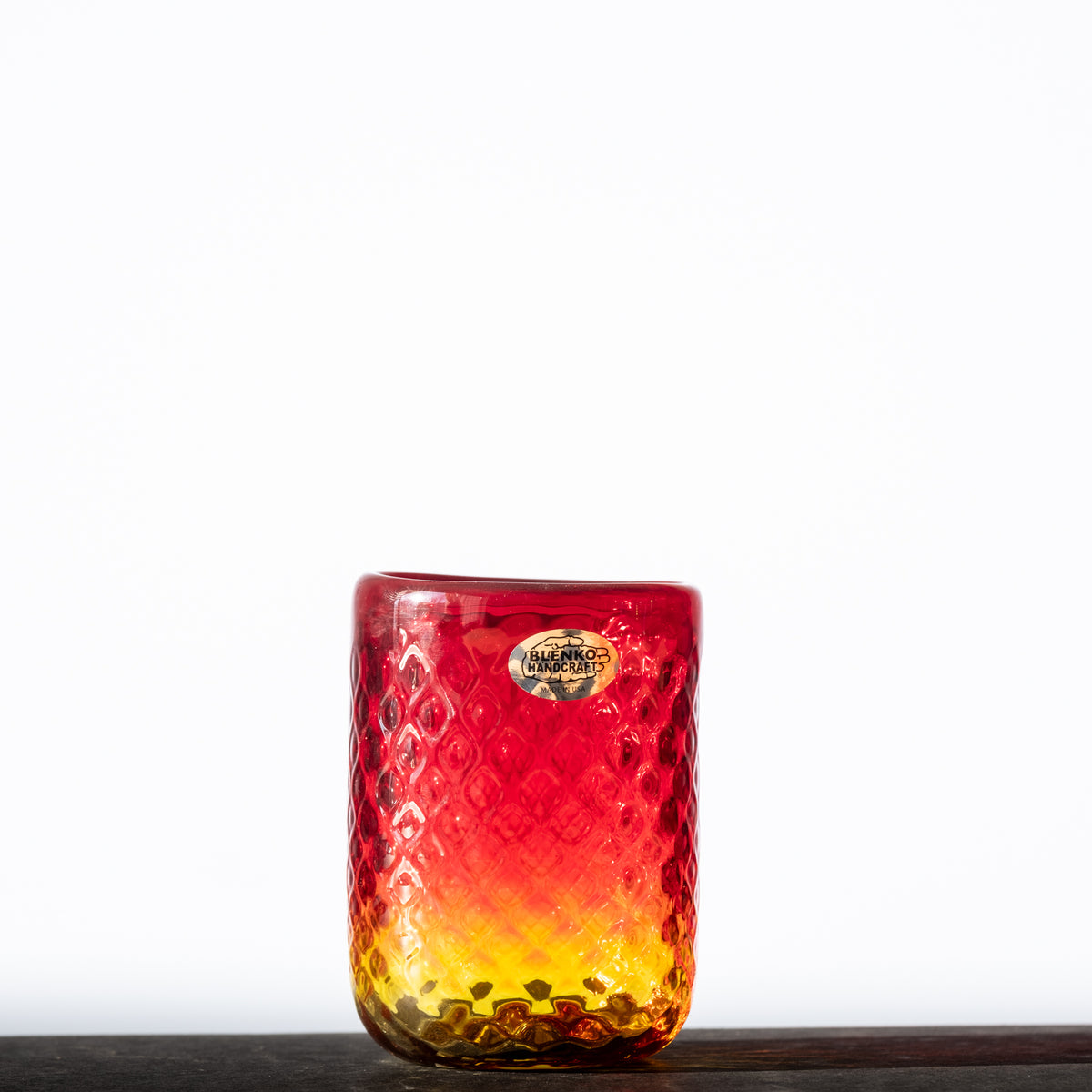 A small, tangerine-colored glass diamond optic tumbler with a textured pattern, displaying a blend of red and orange hues.