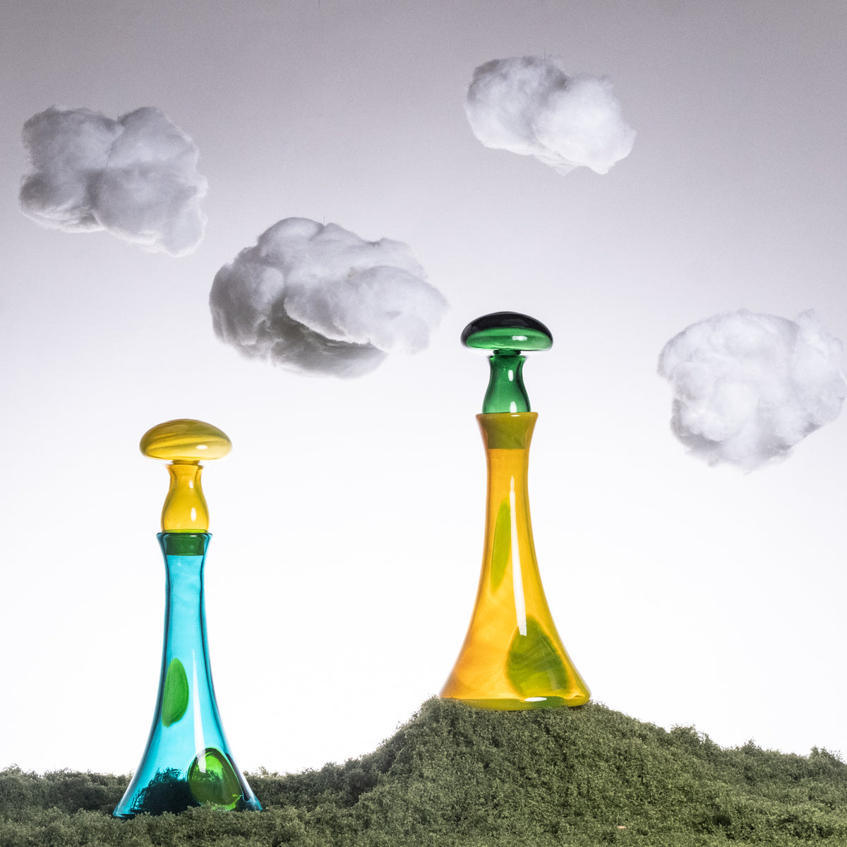 Two colorful glass bottles on a grassy surface with cloud-like shapes in the background.