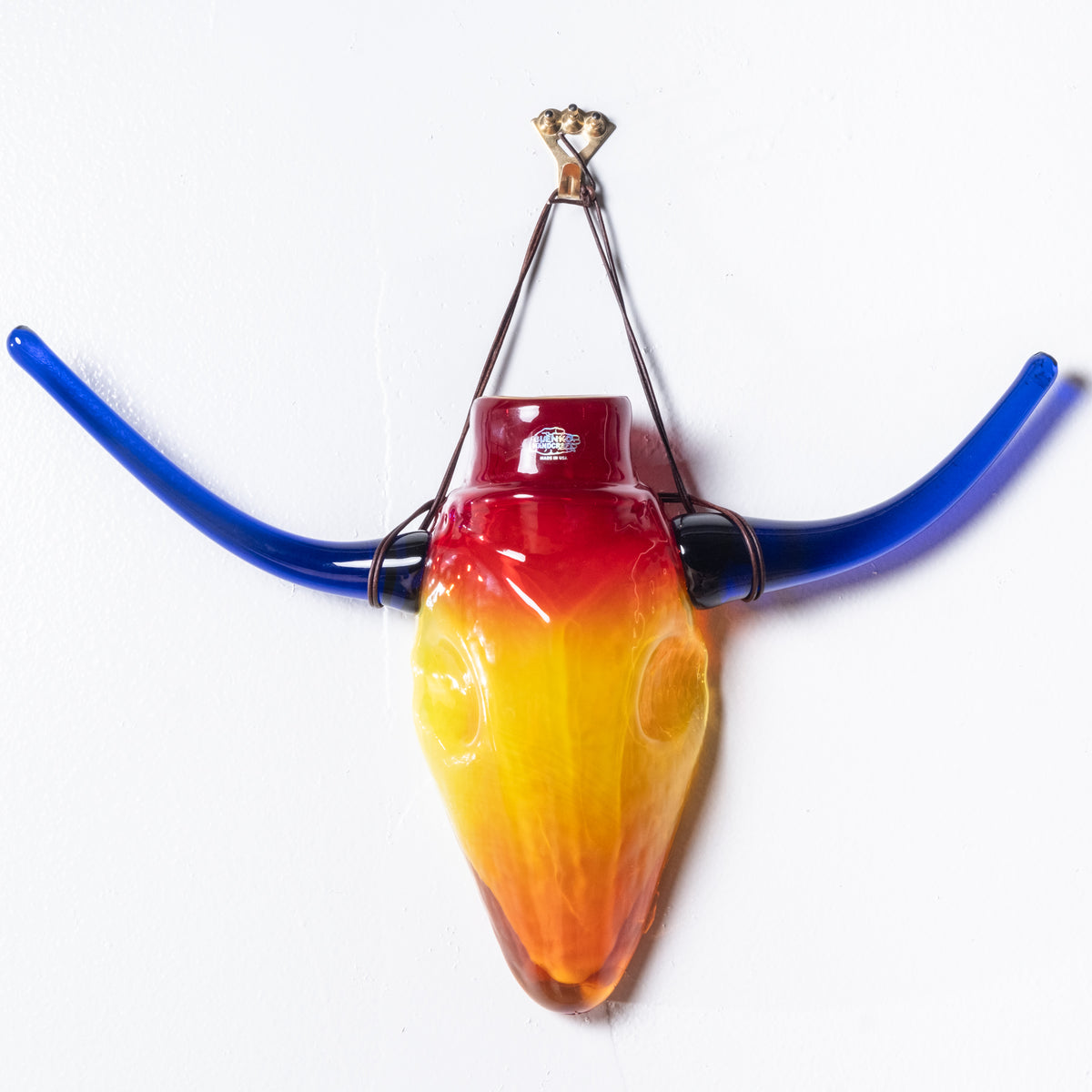 Orange and red glass steer head with blue horns hanging from a white wall.