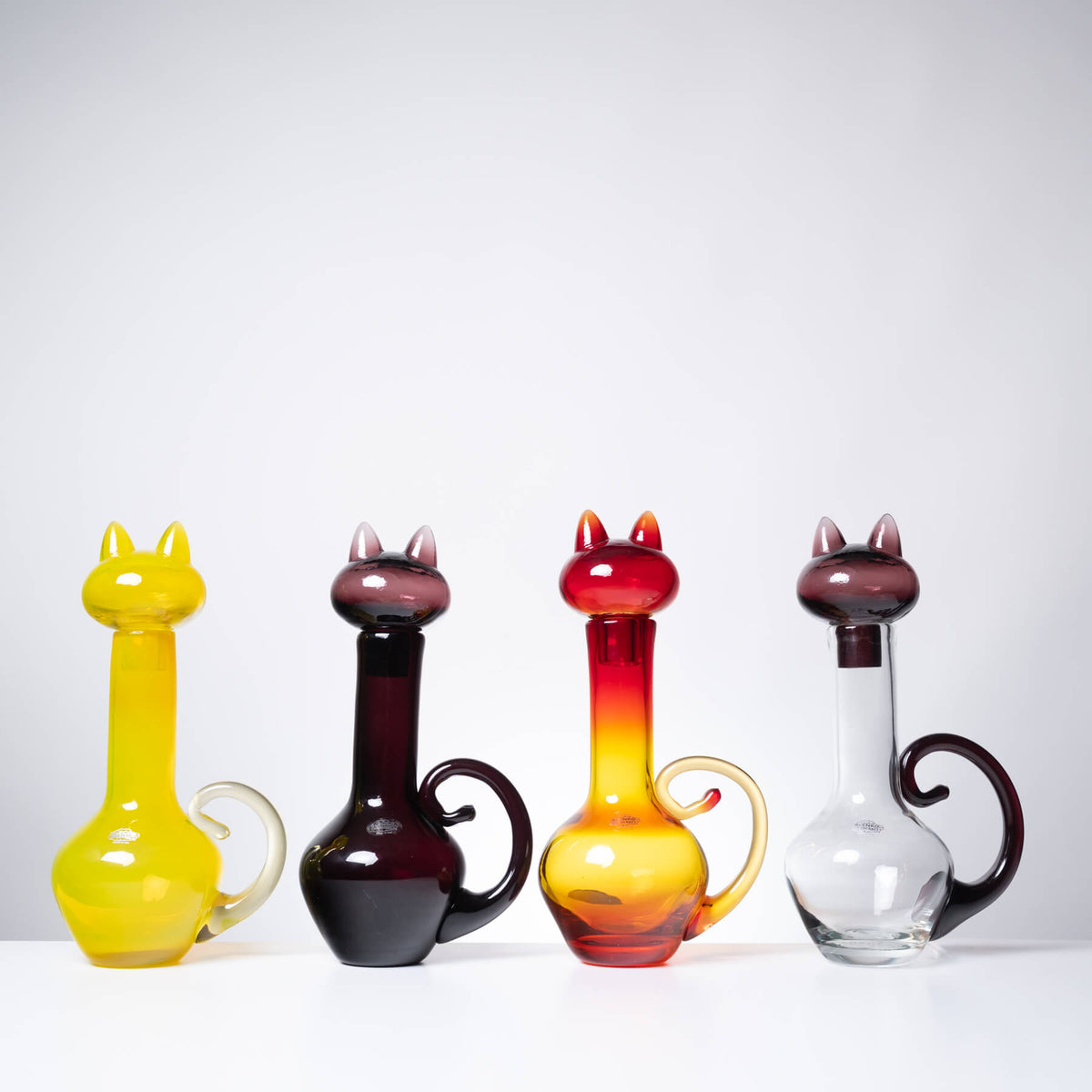 Four cat-shaped glass decanters lined up on a white surface against a light gray background. From left to right, the decanters are yellow with a clear handle, dark burgundy with a dark handle, red and orange gradient with a gold handle, and clear glass with a dark burgundy handle.