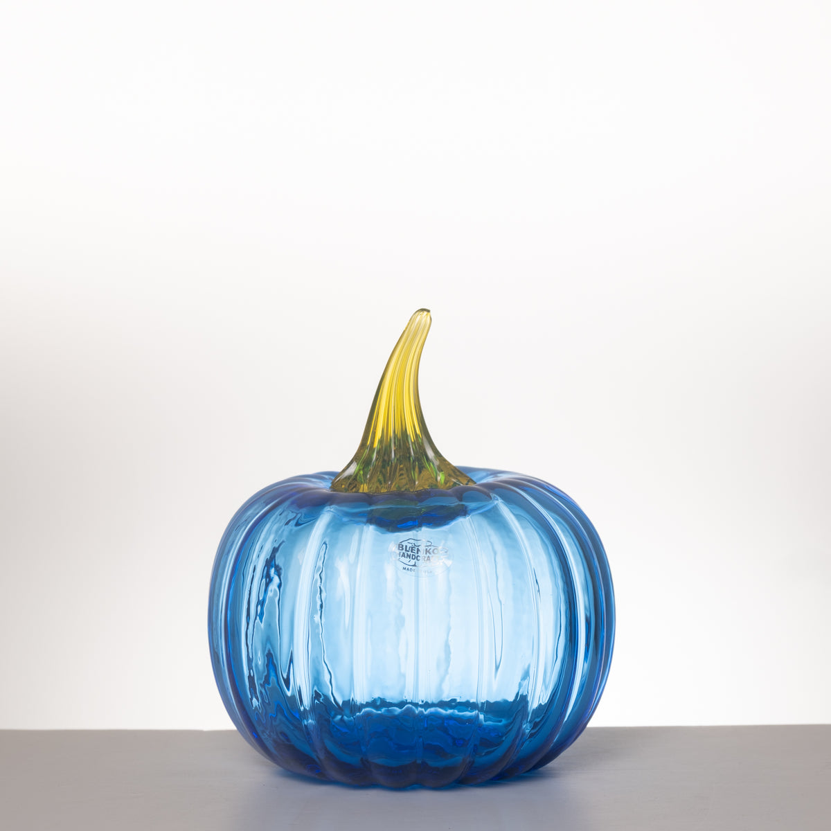 A ribbed glass pumpkin vase in translucent blue with a yellow-green stem, photographed on a neutral gray surface against a white background.