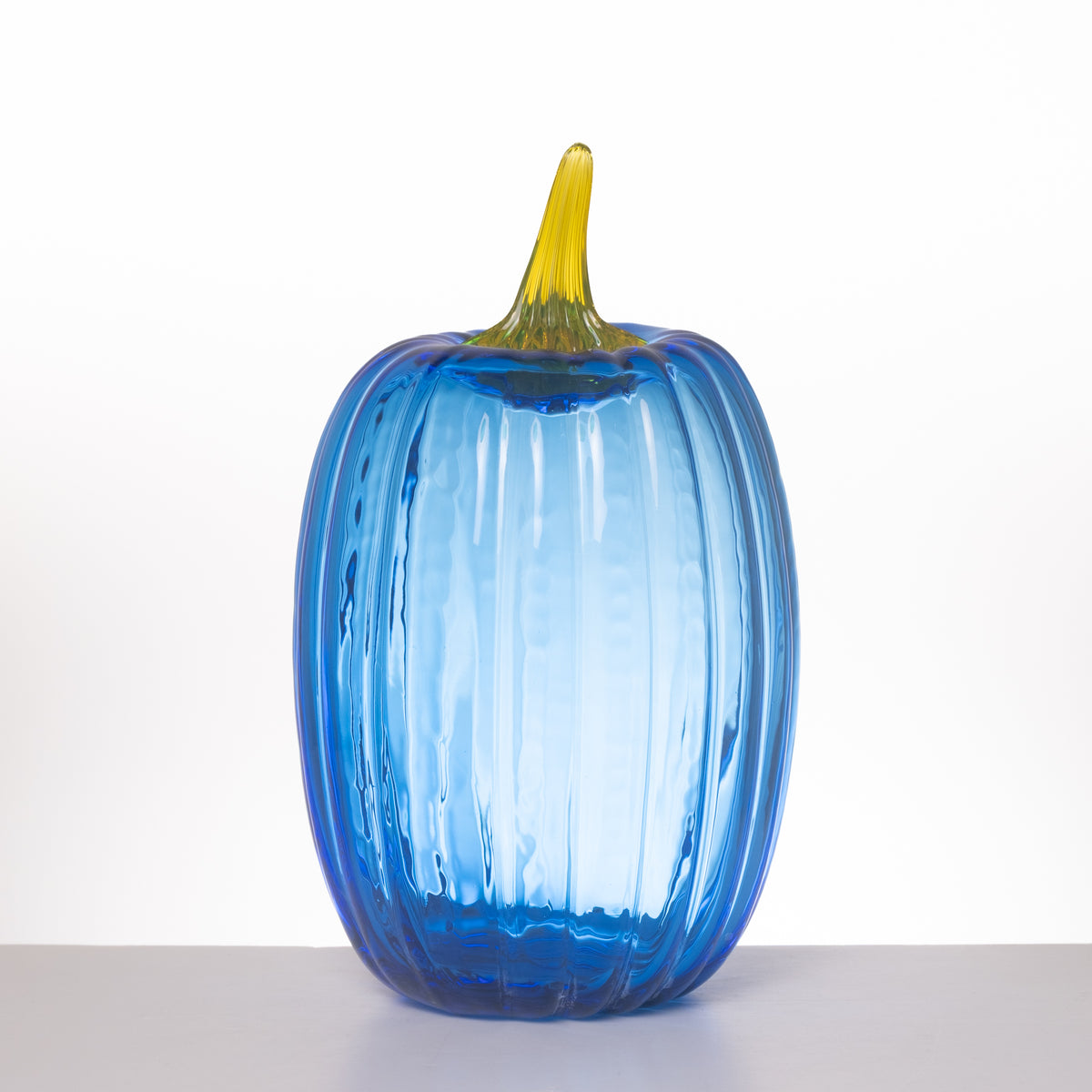 A ribbed glass pumpkin vase in translucent blue with a yellow stem, standing on a light gray surface against a white background.