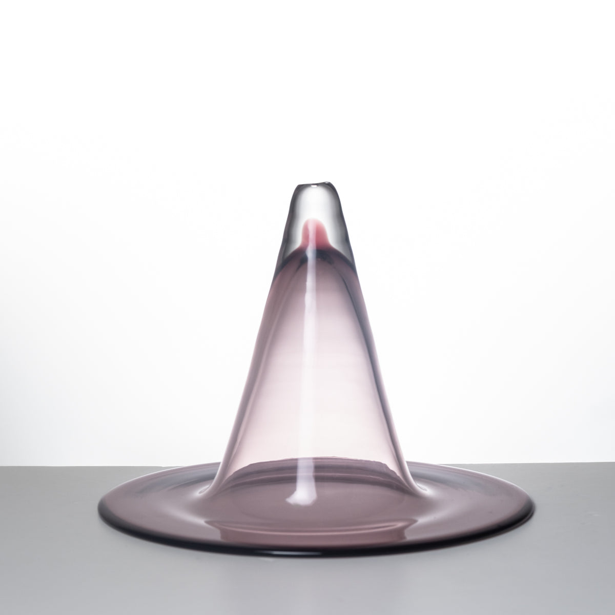 A translucent smoky purple glass sculpture with a wide circular base and a tapered top, photographed on a neutral gray surface against a white background.