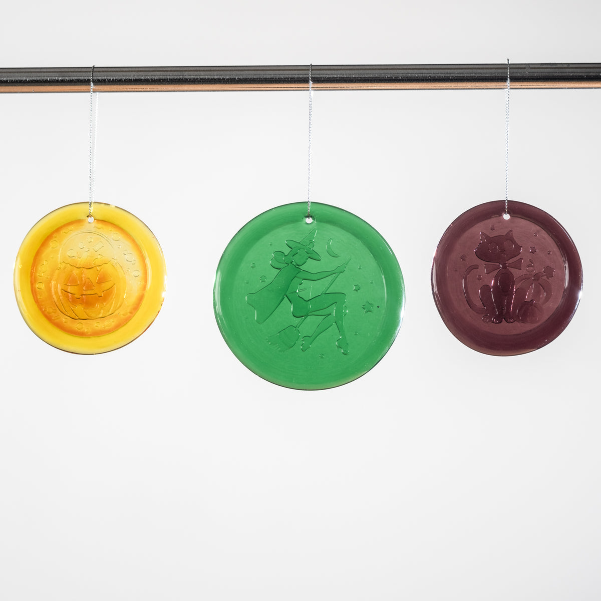 Three round hanging glass ornaments in yellow, green, and dark purple, each embossed with a seasonal design, suspended from a black rod against a white background.