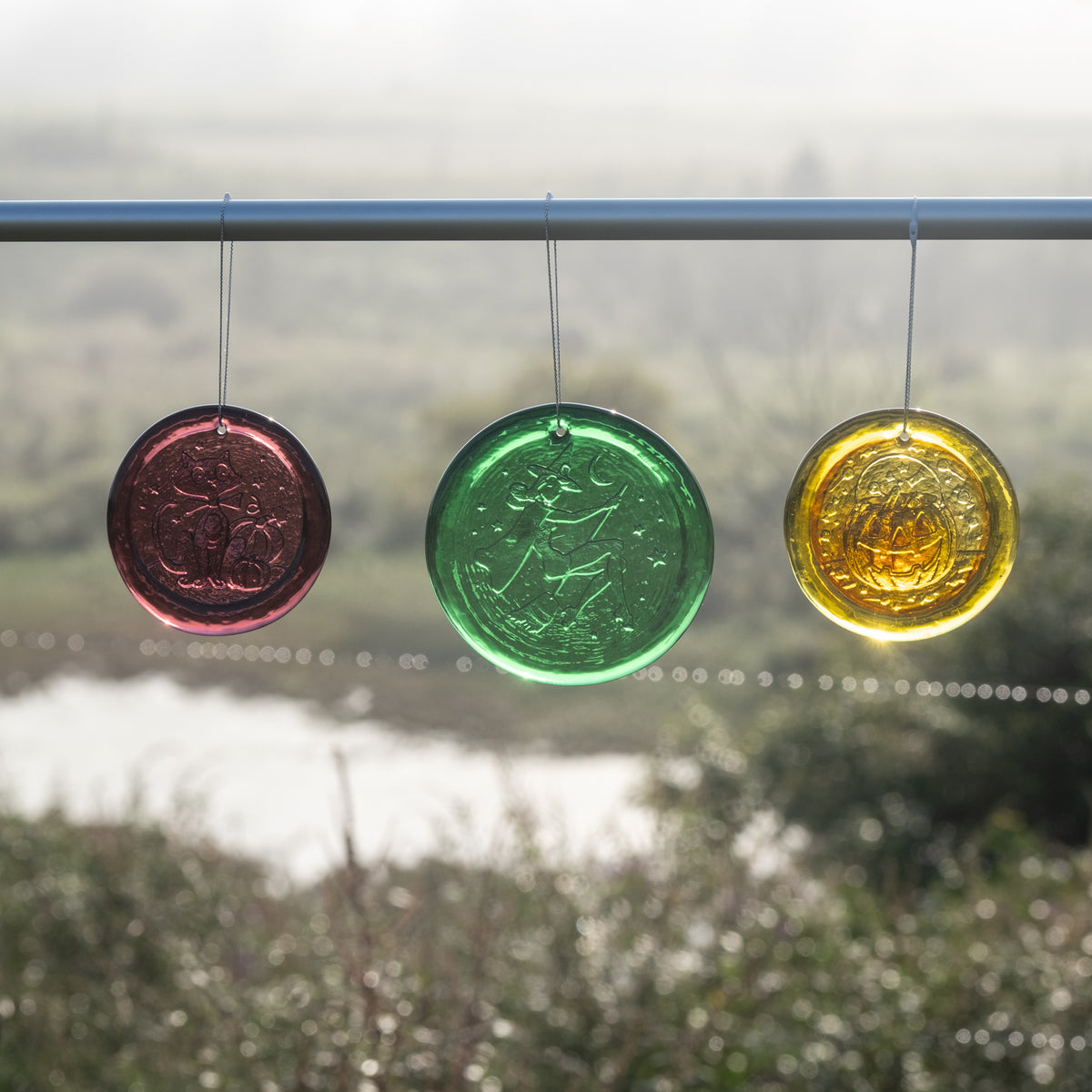 Three circular hanging glass ornaments in red, green, and yellow are suspended from a metal rod outdoors, with a blurred landscape in the background.