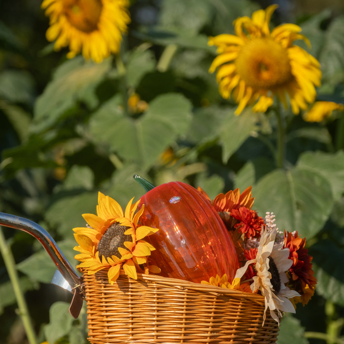 A wicker basket filled with a glossy orange-red decorative pumpkin and sunflowers, set against a blurred background of real sunflowers and green foliage.