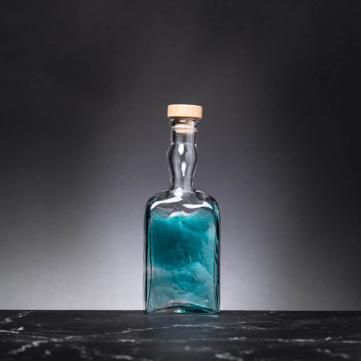 Clear glass bottle with blue liquid and cork lid on a dark surface and gray background