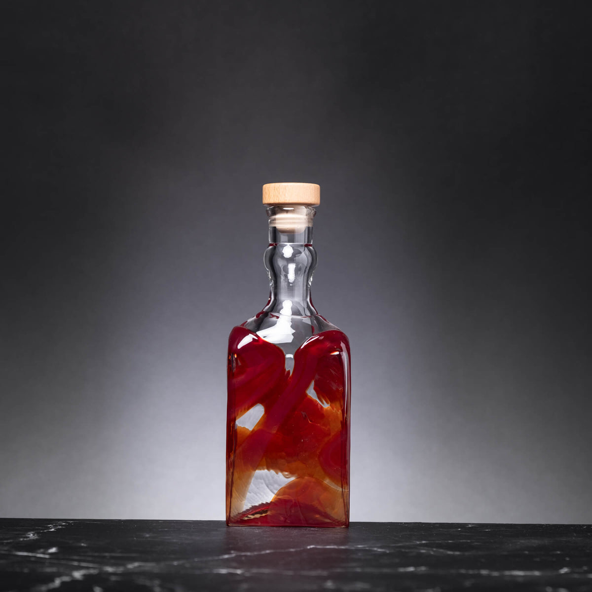 Clear glass bottle with cork lid containing a red liquid on a dark surface and background