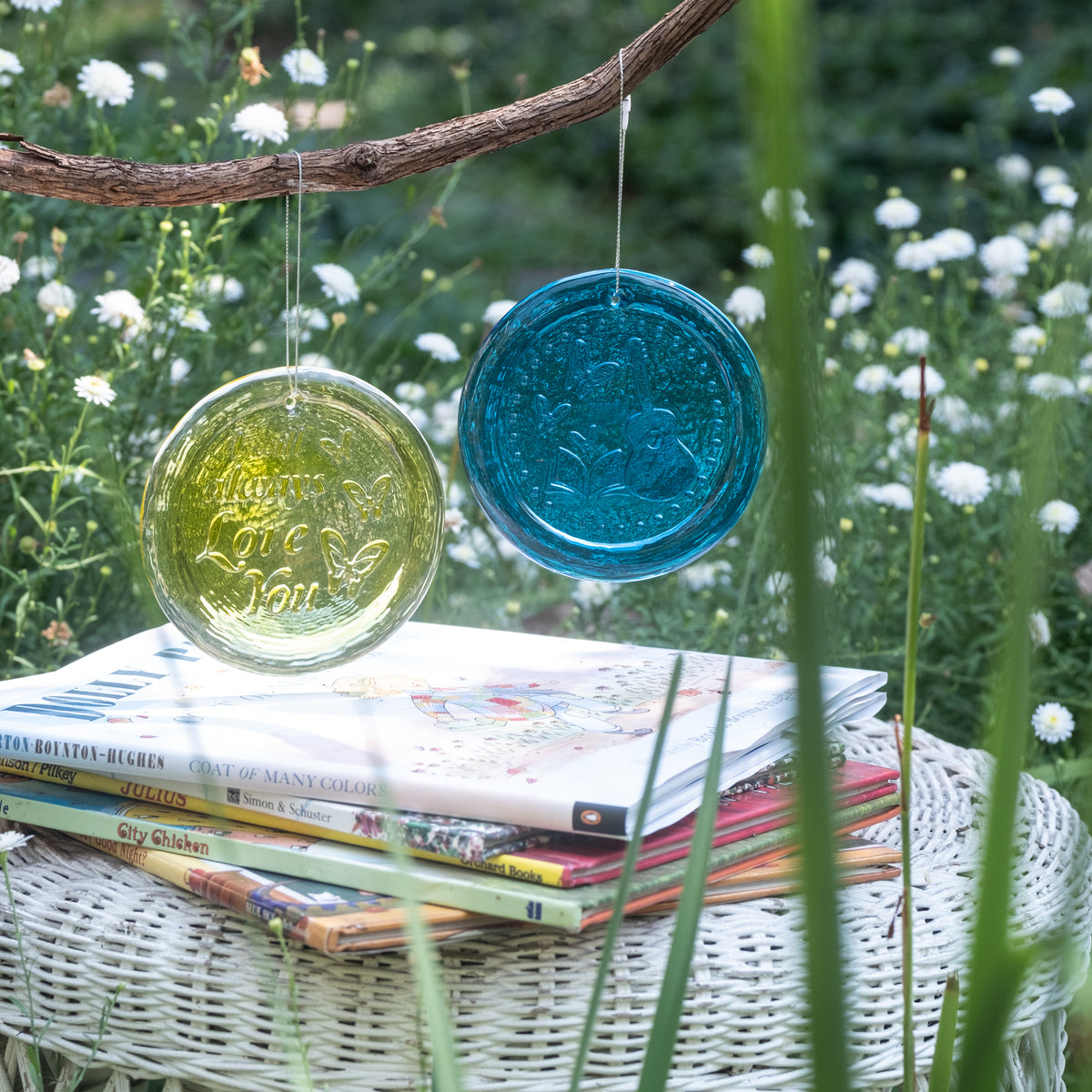 Two round hanging glass ornaments, one translucent yellow-green and one translucent blue, suspended from a tree branch above a white wicker table with stacked books in a garden setting.
