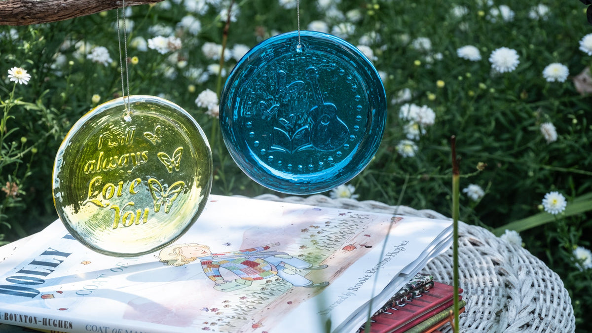 Two decorative glass wind chimes, one translucent yellow-green and one opaque teal blue, hanging outdoors on a stack of books in a garden setting.