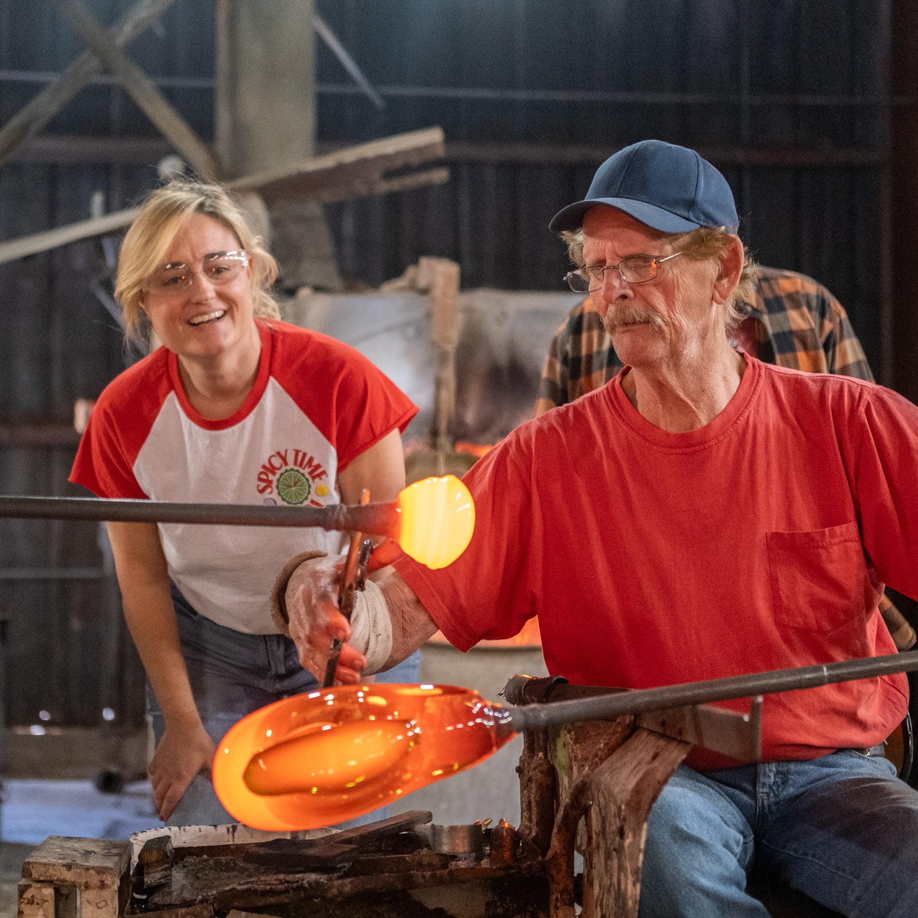 Two people working with molten glass in a workshop setting.