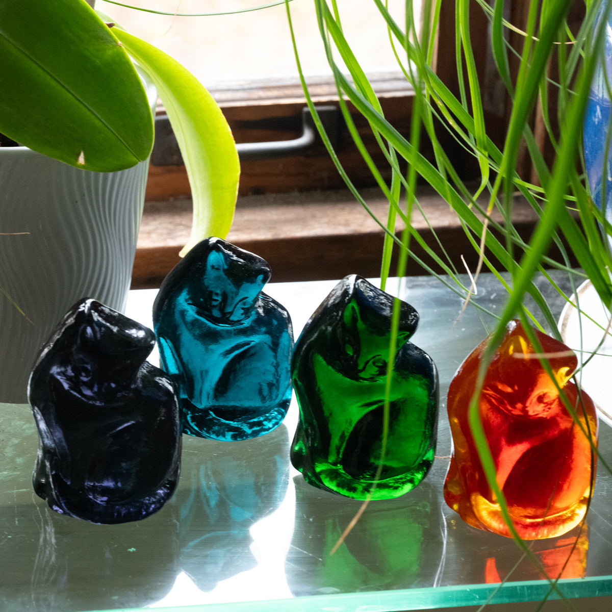 Handcrafted glass cat figurines with a flat back, displayed on a table with plants
