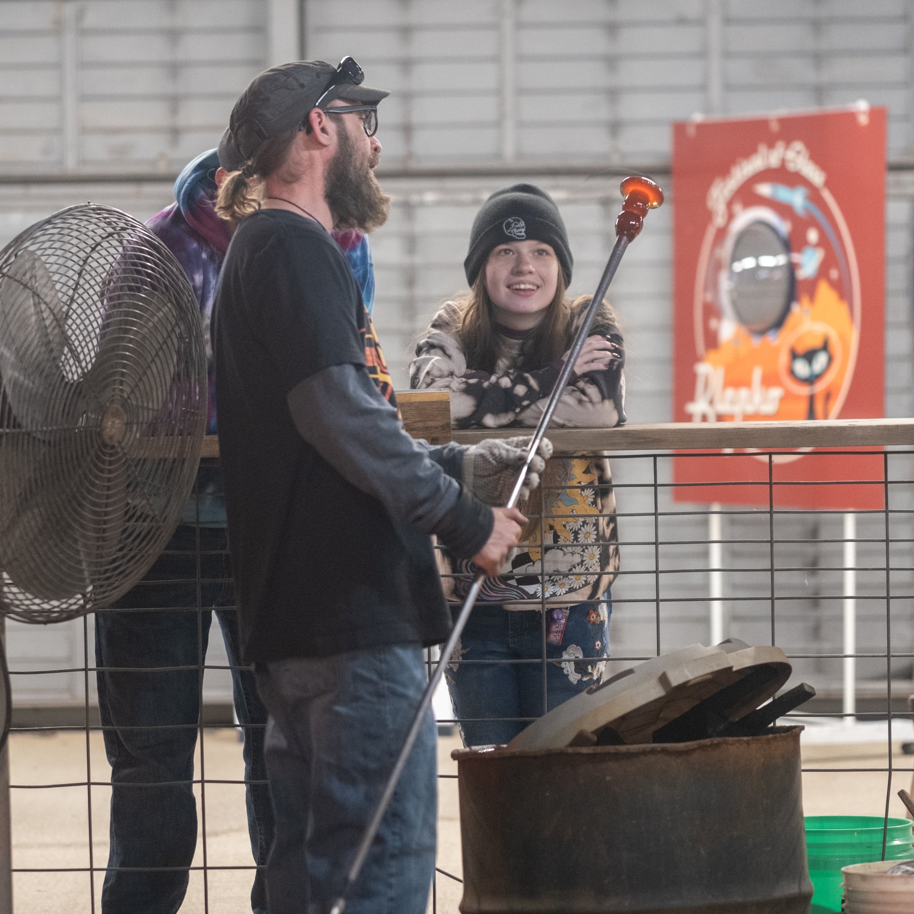 Two people interacting with a metal object near a fan and a colorful banner in the background.
