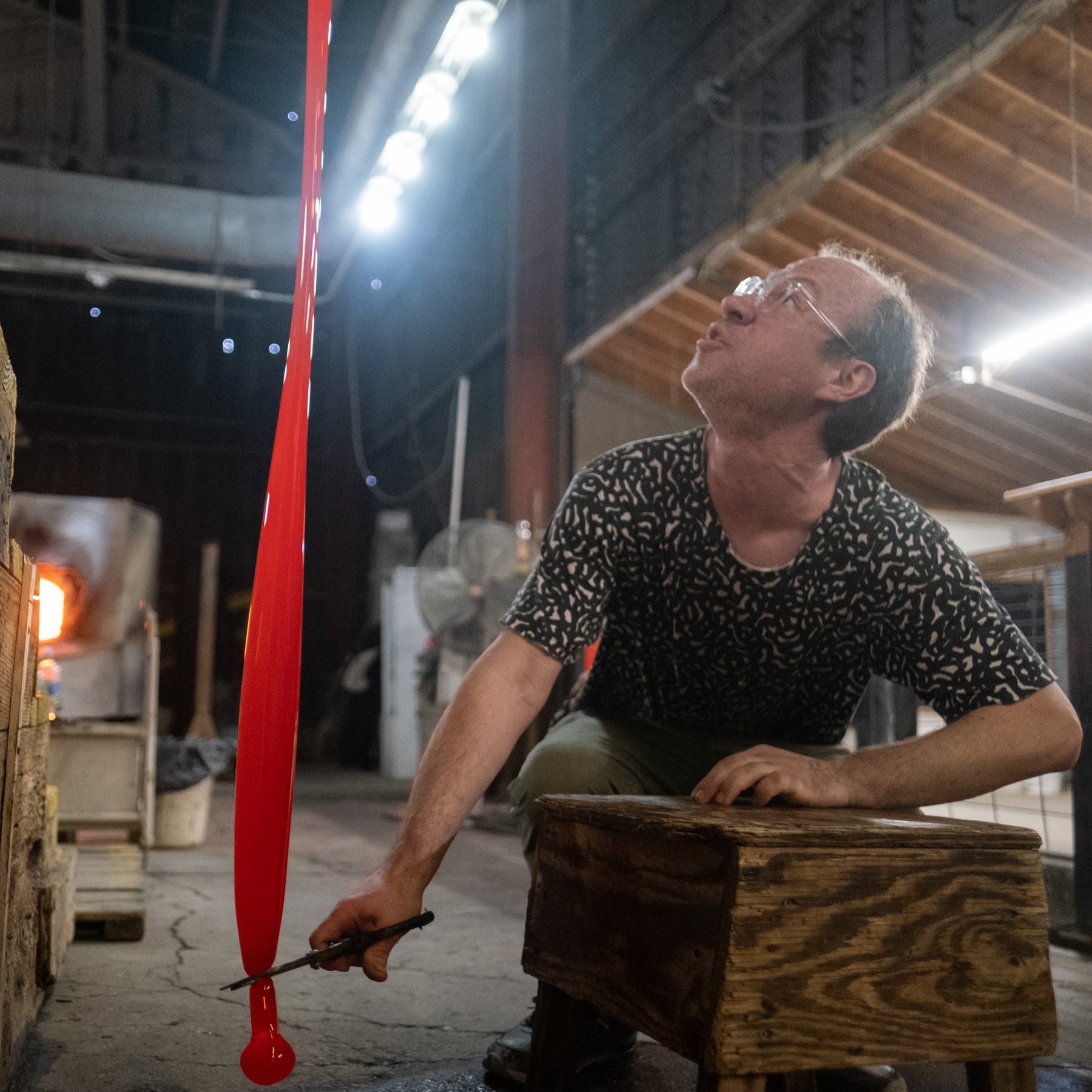 Man working with glass in a workshop, holding a long red glass object.