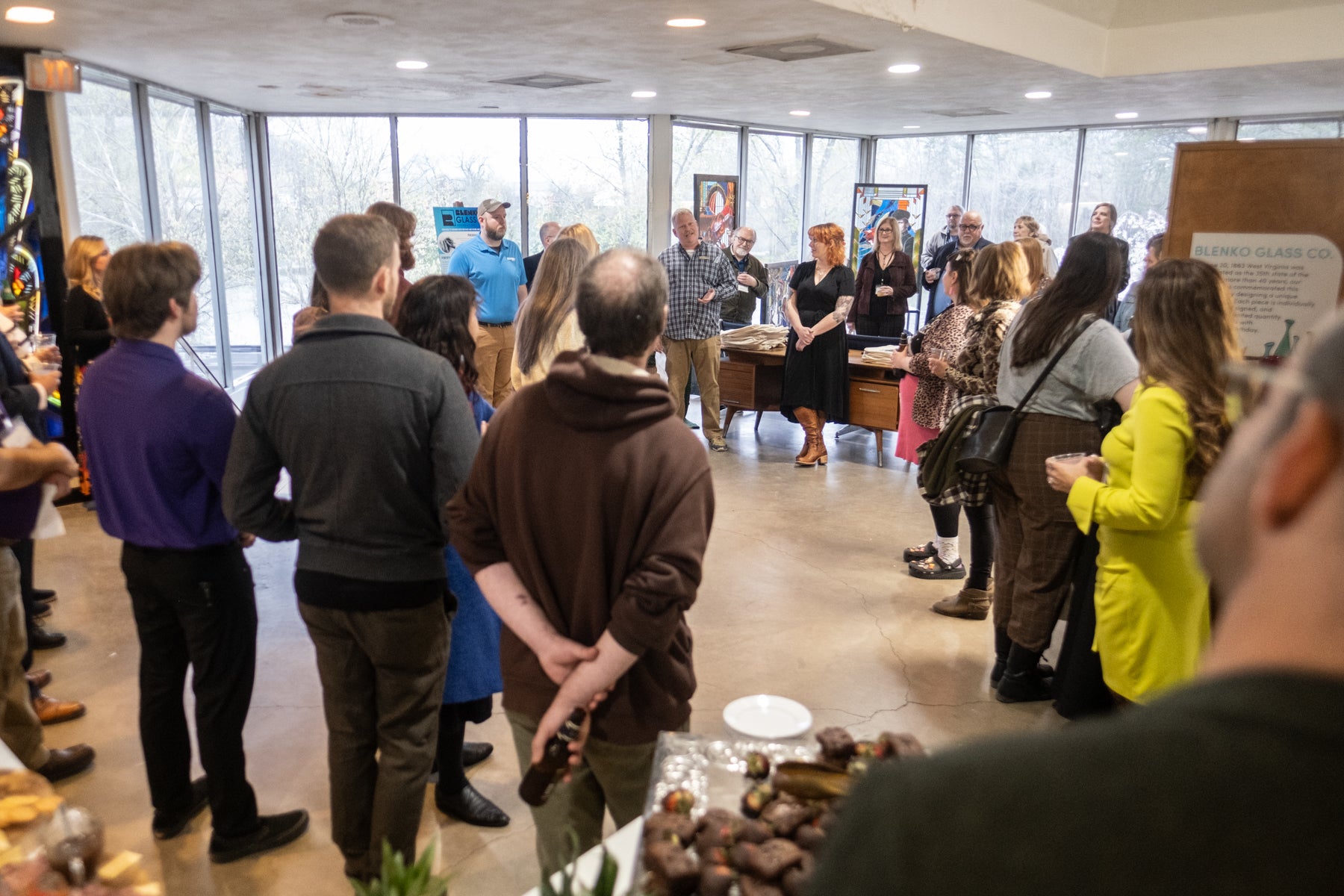 Group of people gathered in a room with large windows, possibly at an event or exhibition.