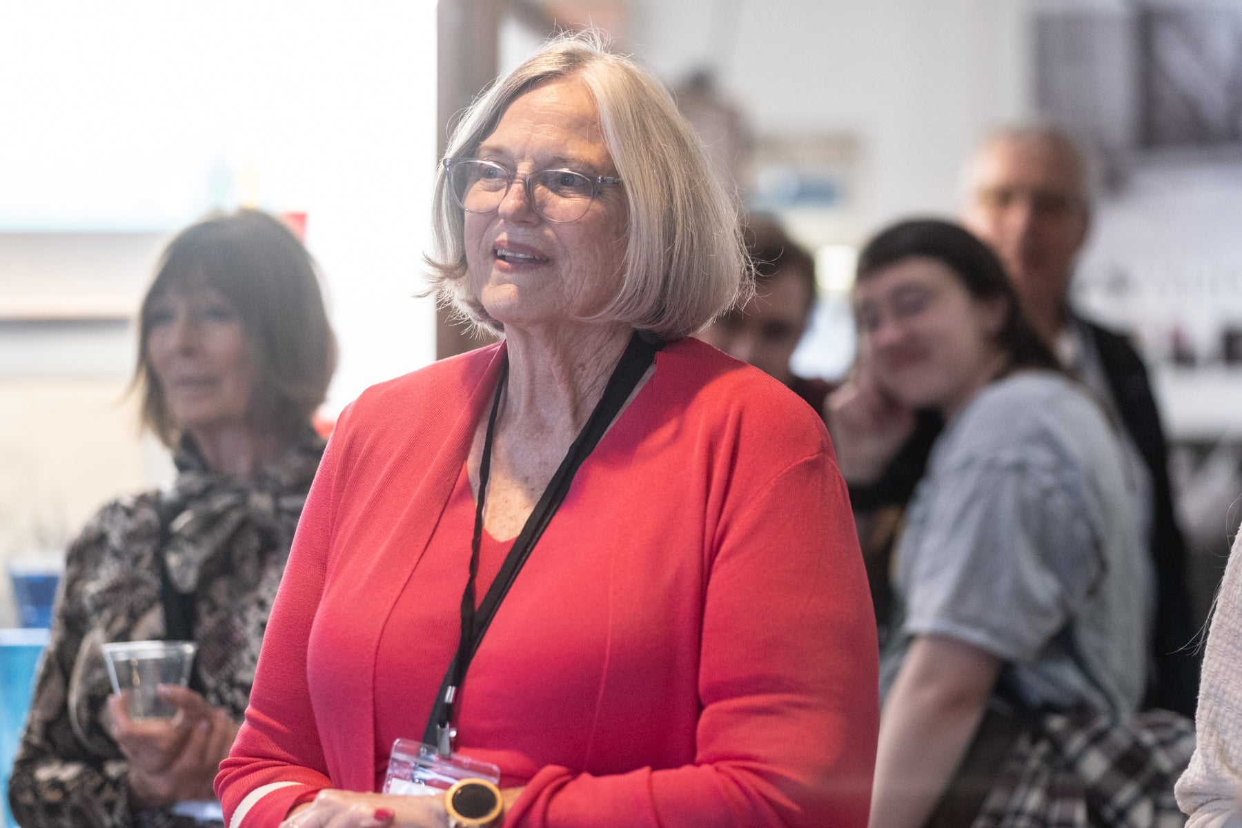 Woman in a red sweater with a lanyard, standing in an indoor setting with blurred people in the background.