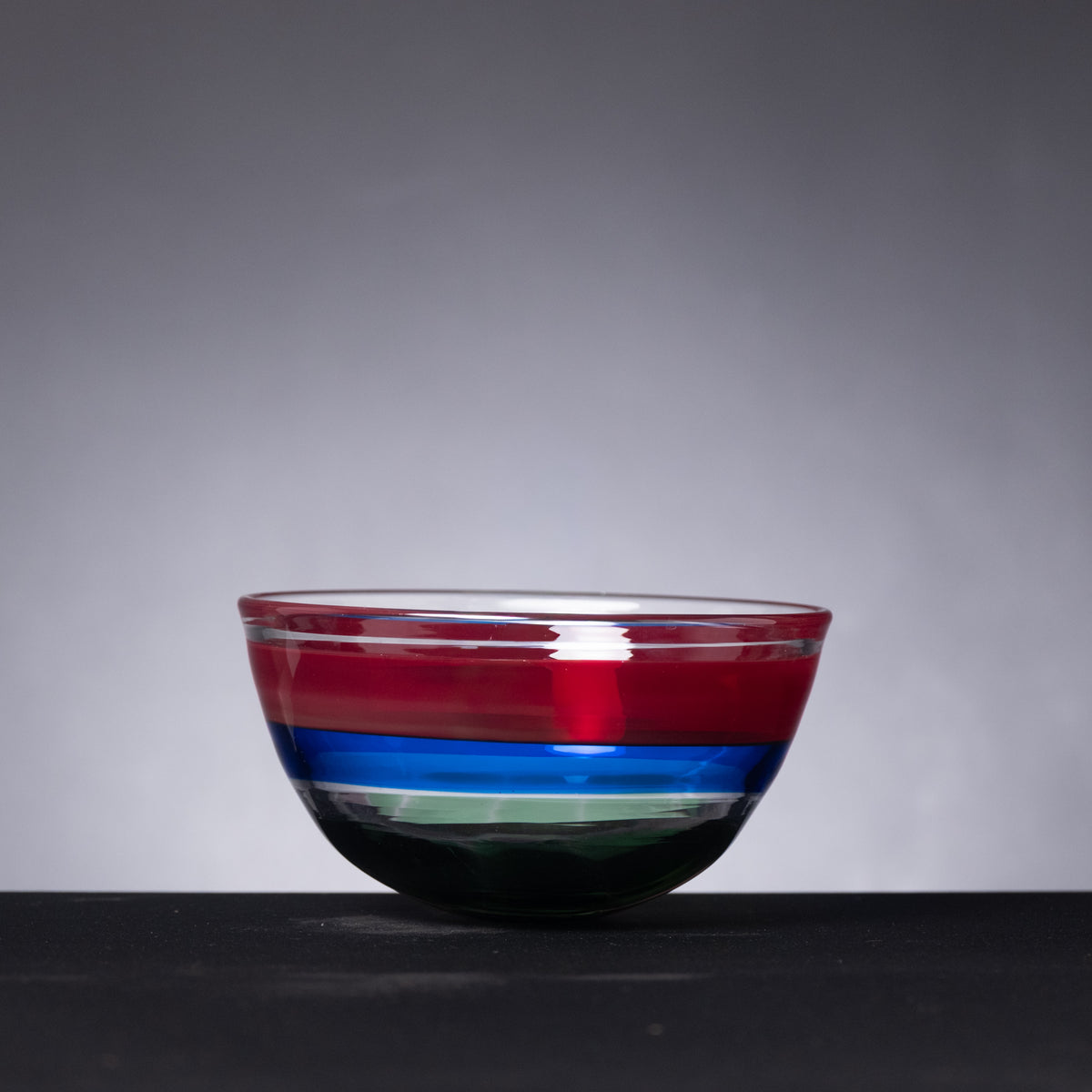 Colorful glass bowl with red, blue, and green stripes on a dark surface and gray background