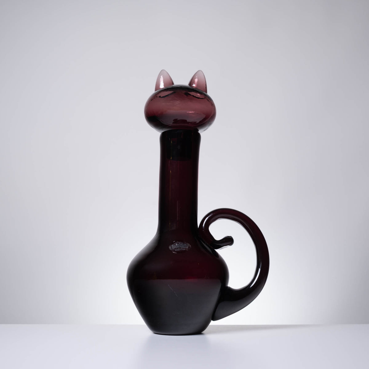 A dark burgundy glass decanter shaped like a cat, featuring pointed ears on the neck, a curved handle, and a glossy finish, set against a neutral gray background.