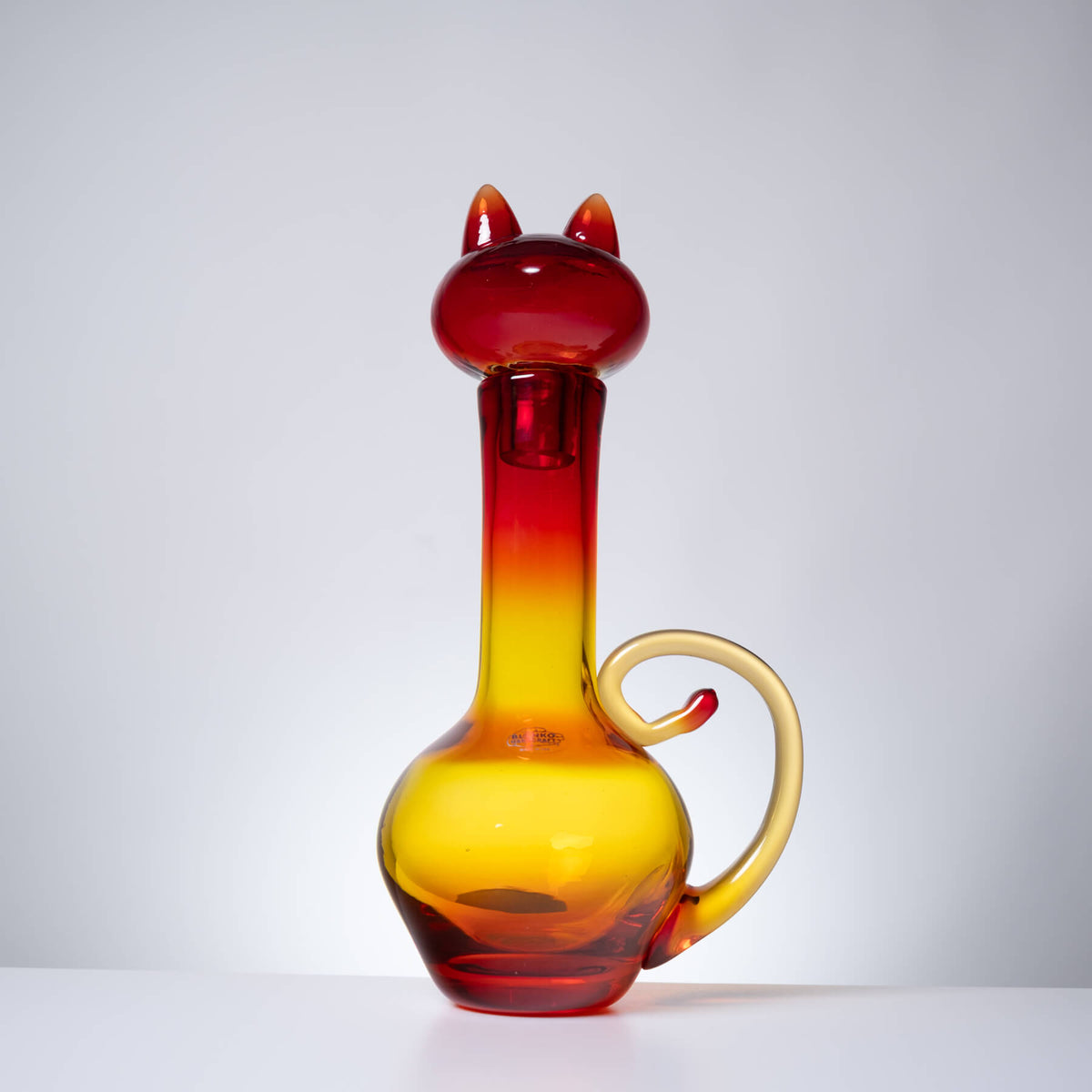 A translucent glass carafe shaped like a cat, featuring a red and yellow gradient body with a curved handle and cat ears on the neck, set against a plain light gray background.