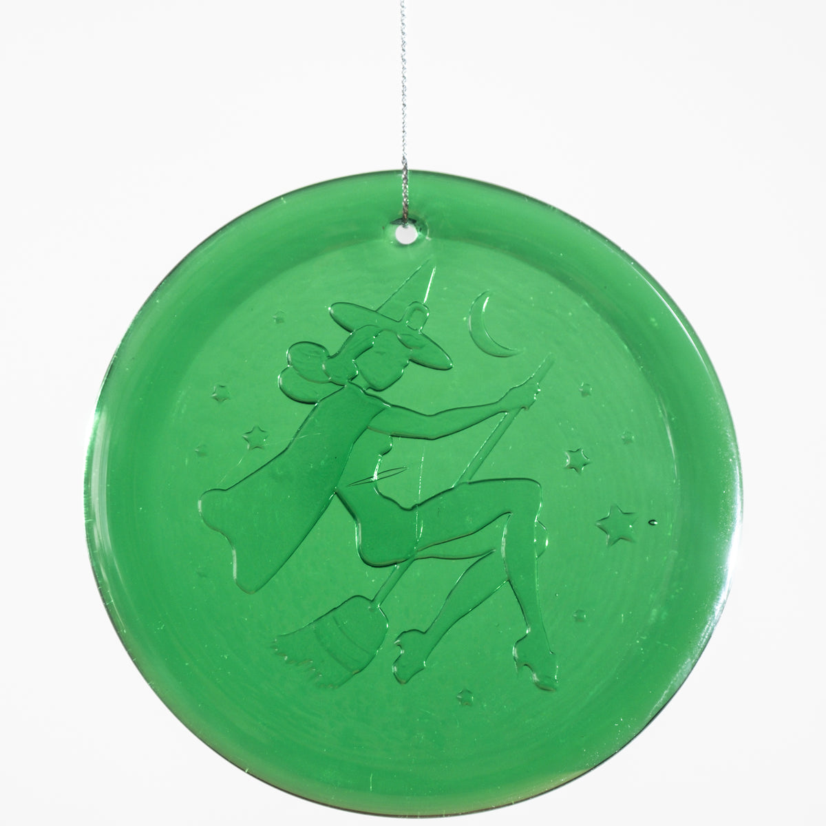 A round green glass hanging ornament featuring an embossed design of a witch riding a broomstick under a crescent moon, suspended by a thin string against a white background.