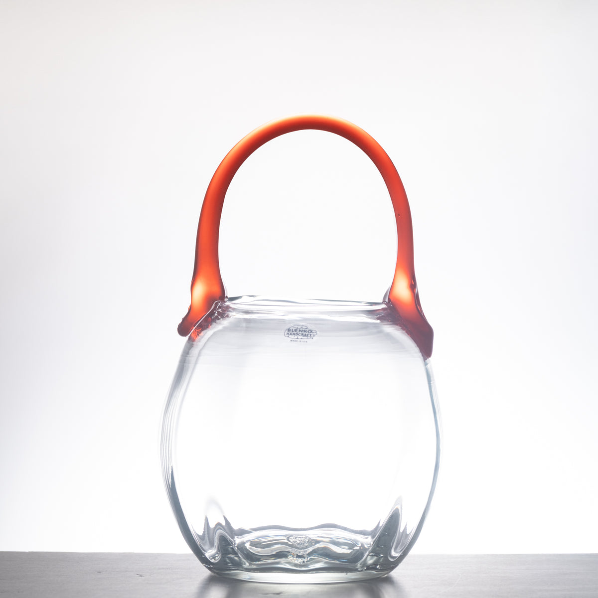 A clear glass handbag with a rounded, fish-shaped body and a vibrant orange handle, shown on a neutral light gray background.