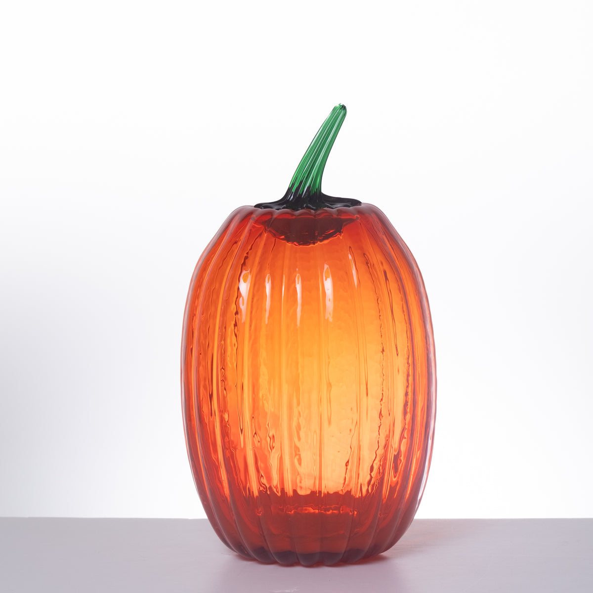 A ribbed glass pumpkin vase with a vibrant orange body and a green stem, set against a plain white background.