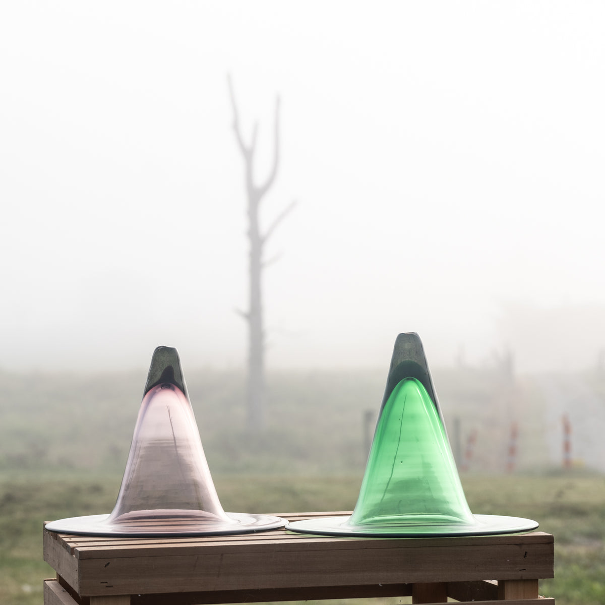 Two translucent cone-shaped glass vessels, one pink-tinted and one green-tinted, displayed side-by-side on a wooden table in a misty outdoor setting.