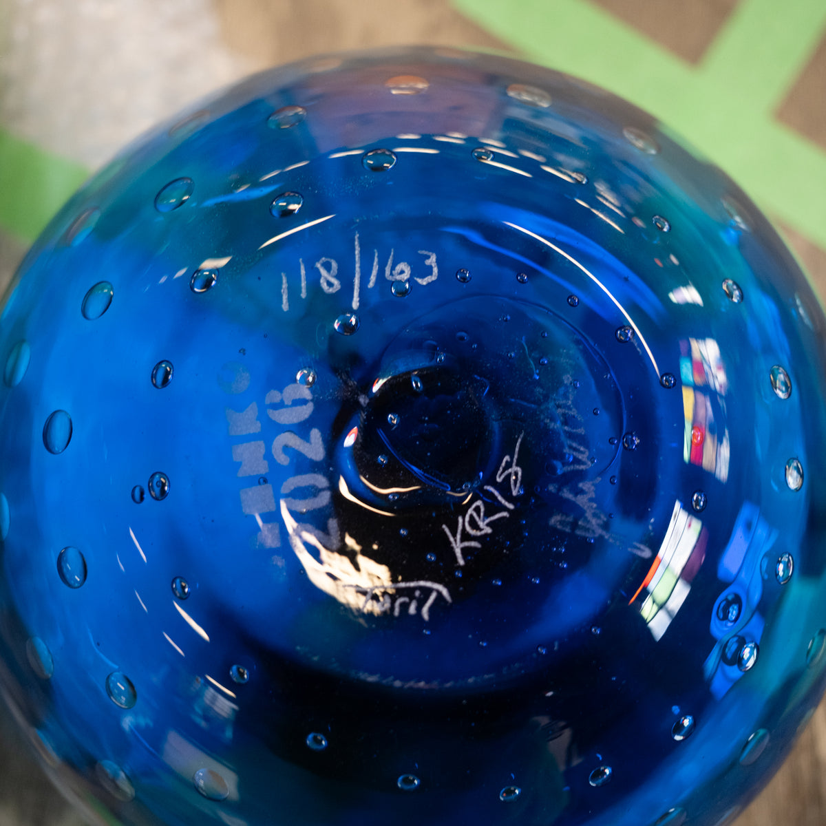 A close-up of a blue glass bowl or vase showing the bottom of the vessel, with white handwritten markings and bubbles in the glass, photographed against a blurred background.