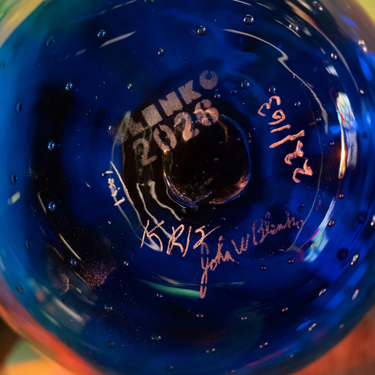 Close-up of the bottom of a deep cobalt blue glass bottle or vase, featuring embossed text reading 'LINKO 2028' and two metallic signatures, one in gold and one in rose gold, along with the date '22/16'.
