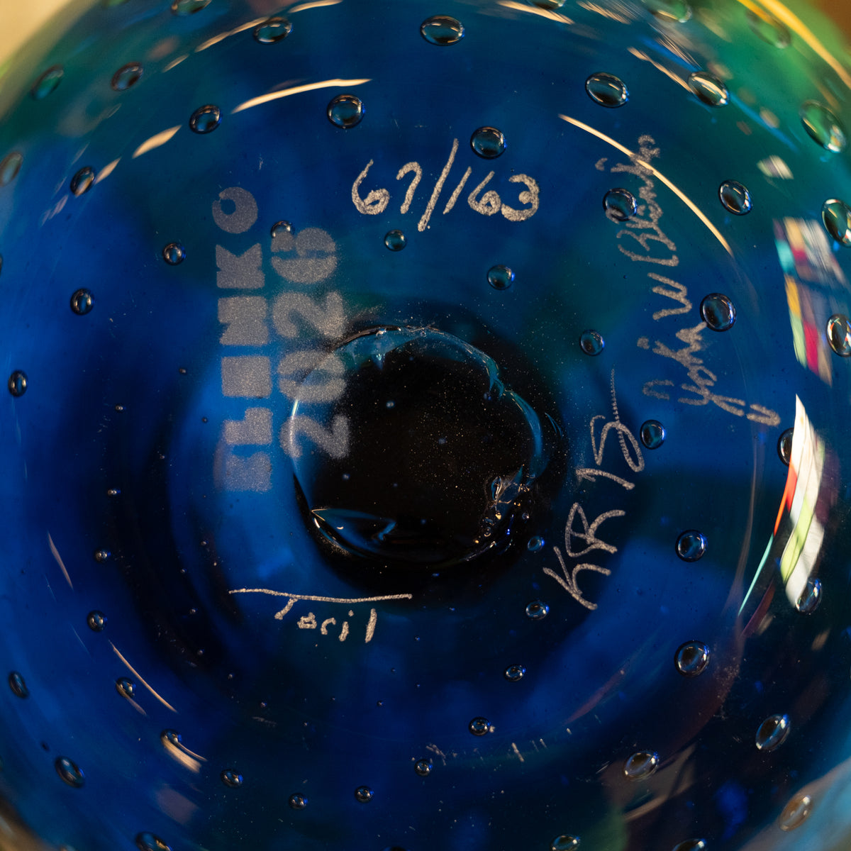 Close-up of a blue glass bowl or vase with a circular center, showing white handwritten markings and signatures on the inside surface.