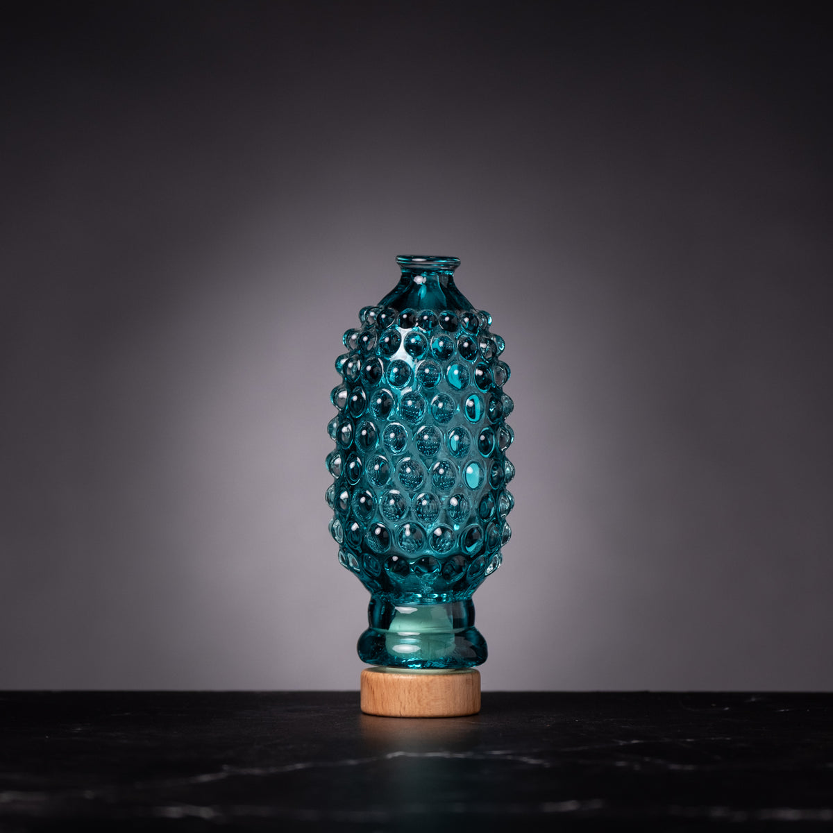 Teal glass vase with wooden base on a dark surface and gray background