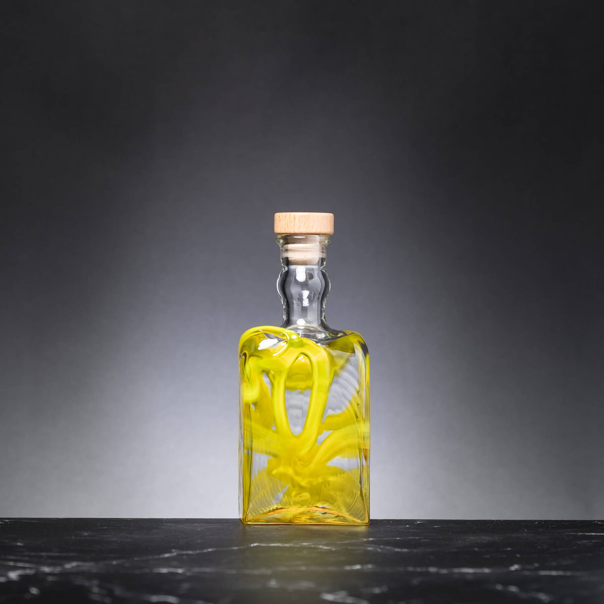 Clear glass bottle with a cork lid containing a yellow liquid on a dark surface and gray background
