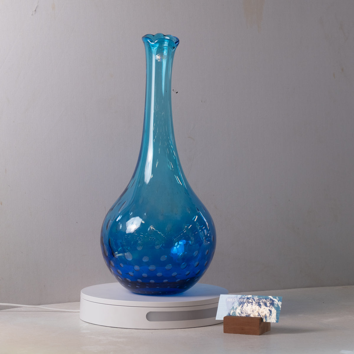 A translucent blue glass vase with a flared neck and dotted pattern near the base, displayed on a white circular stand against a plain light gray wall.