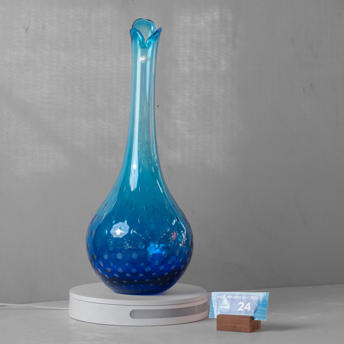 A blue glass decanter with a flared rim and dotted textured pattern, resting on a white circular scale with a small weight card next to it, set against a gray wall.