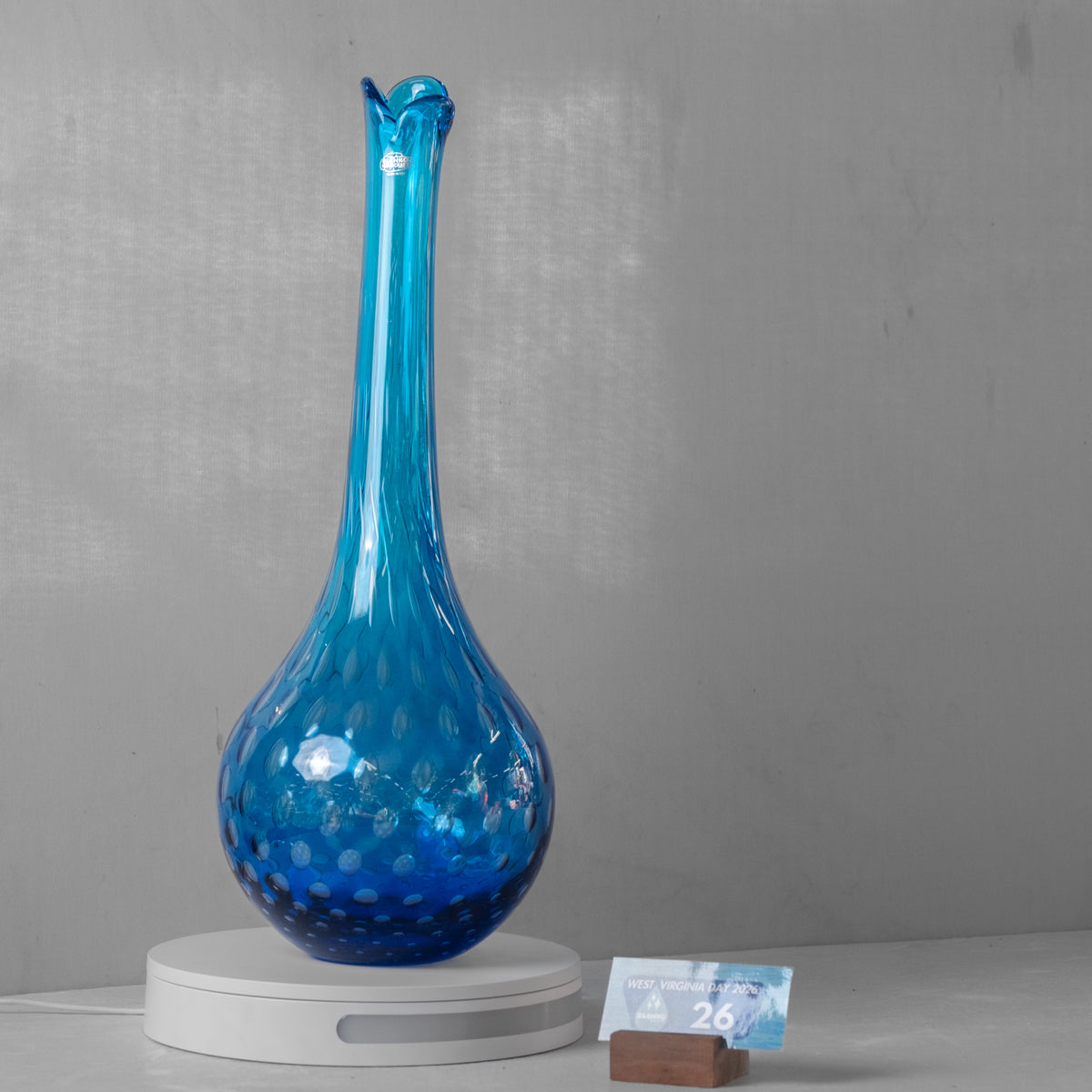A tall, bulb-shaped blue glass vase with a narrow neck and a hammered texture, displayed on a white circular scale with a small price tag in front, against a gray wall.